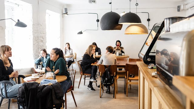 Group of multiracial friends in casual wear sitting at wooden tables in cozy cafeteria near cash box and big lamps while conversing